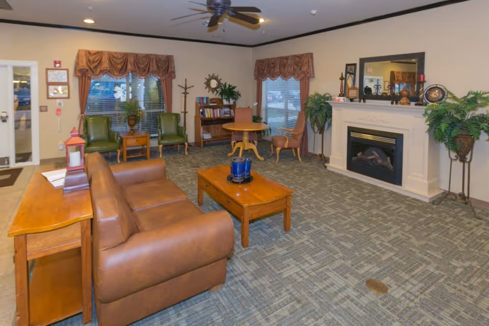 A cozy living room area in EastView HealthCare & Rehabilitation Center featuring a brown leather sofa, wooden coffee table with a blue candle holder, two green armchairs with a small wooden side table between them, a round wooden table with two chairs, a fireplace with a large mirror above it, and several potted plants. The room has carpeted flooring and windows with brown valances.