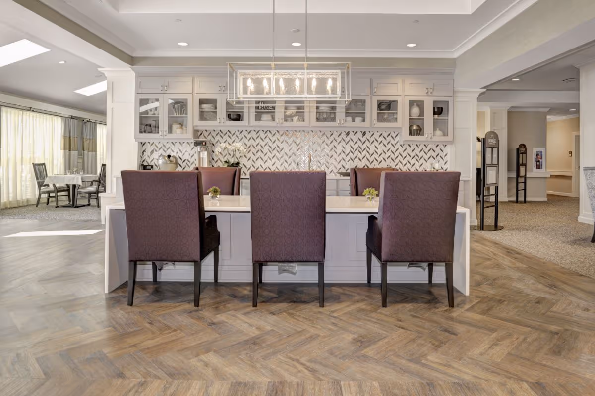 Bright communal kitchen and dining area with a central island, three upholstered bar chairs, upper cabinets and a patterned tile backsplash.