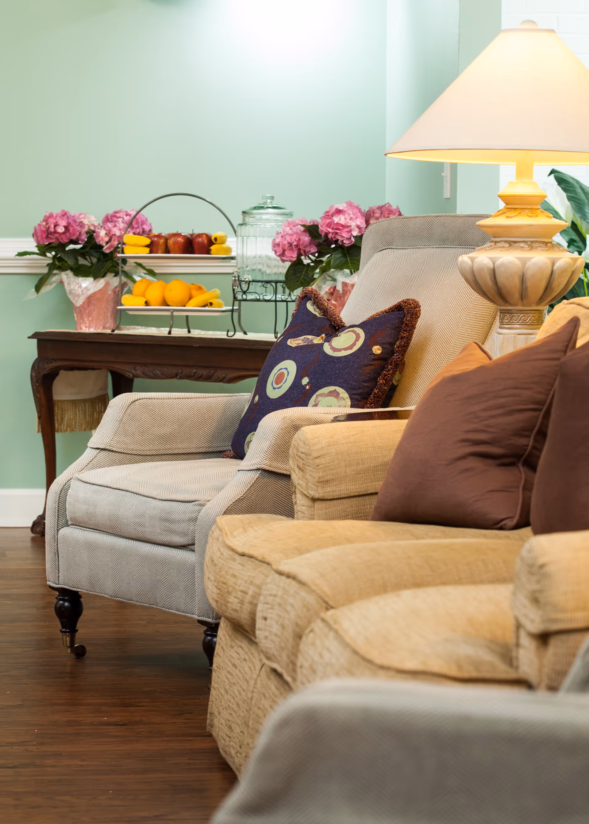 Cozy living room area with beige and light gray upholstered armchairs adorned with decorative pillows. A wooden side table behind the chairs holds a tiered fruit stand with apples and oranges, a glass jar, and two pink flower arrangements. A classic table lamp with a beige shade is also visible, casting warm light in the room with light green walls and wooden flooring.