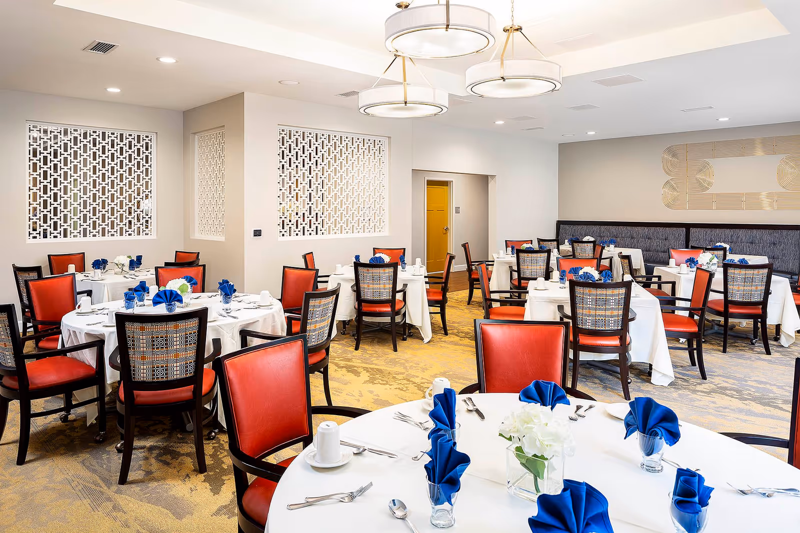 A bright and elegant dining room with multiple round tables covered in white tablecloths, each set with blue folded napkins, white cups, and silverware. The chairs have red cushioned seats and patterned backs. The room features modern light fixtures hanging from the ceiling and decorative wall panels.