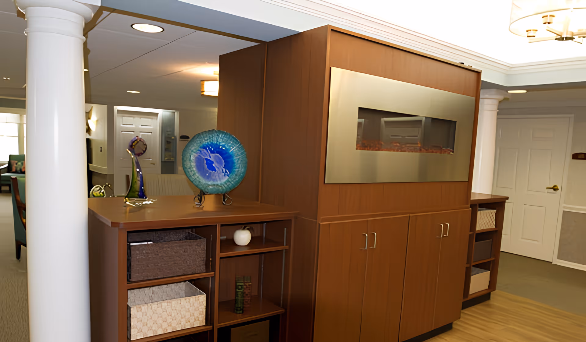 Interior view of a senior living facility featuring a wooden cabinet with an electric fireplace insert. On top of the cabinet are decorative glass art pieces, including a blue circular plate and a purple sculpture. The room has white columns, recessed lighting, and a door in the background.