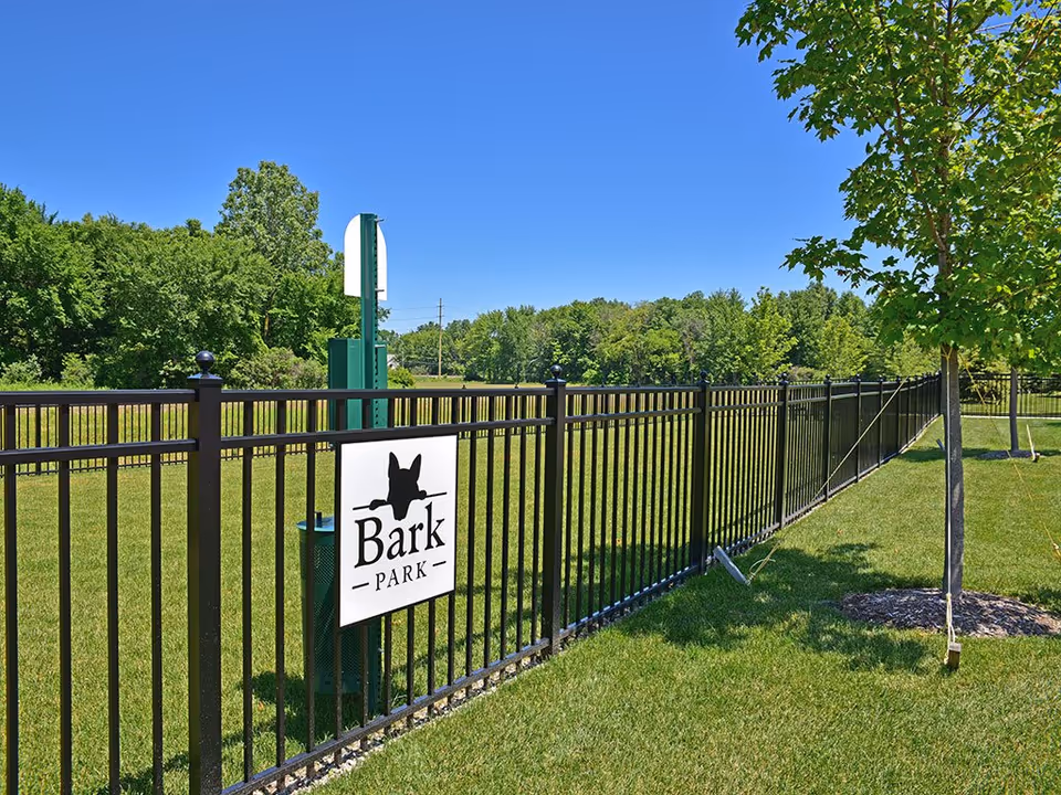 Fenced grassy dog park area with a "Bark Park" sign, trees, and a clear blue sky.