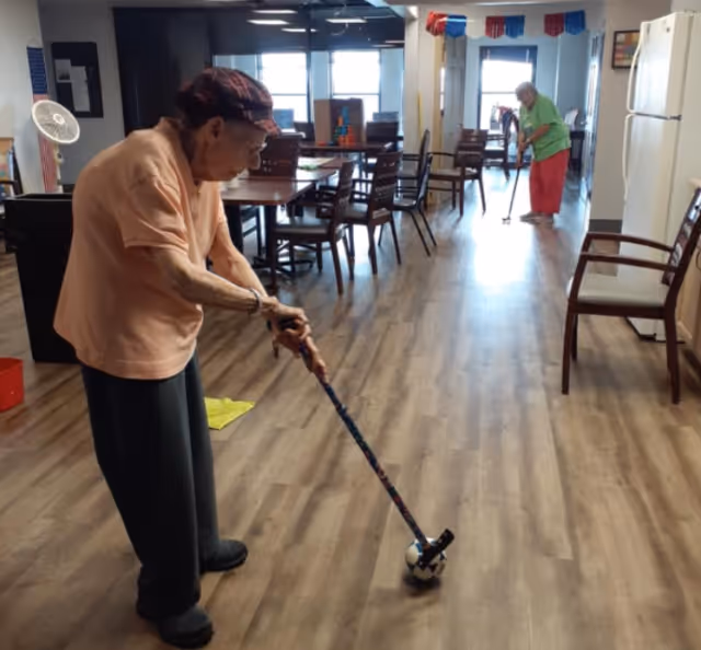 Two elderly individuals playing an indoor game resembling floor hockey in a common area with wooden flooring, tables, and chairs. One person in the foreground is holding a stick with a ball, while another person in the background is also holding a stick. The room is well-lit with windows and has a refrigerator and chairs along the walls.