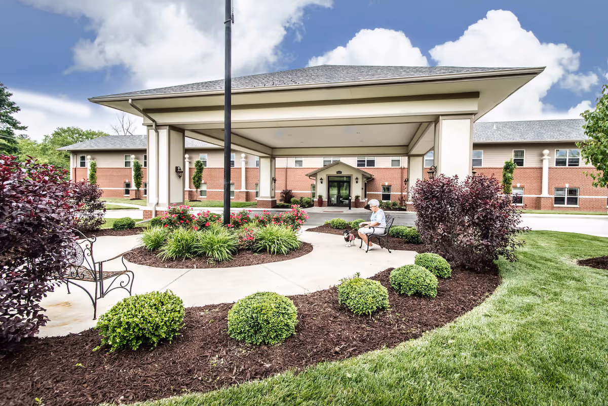 Entrance of Cedarhurst of Columbia senior living facility with a covered drop-off area, landscaped garden beds with bushes and flowers, and a person sitting on a bench with a small dog.