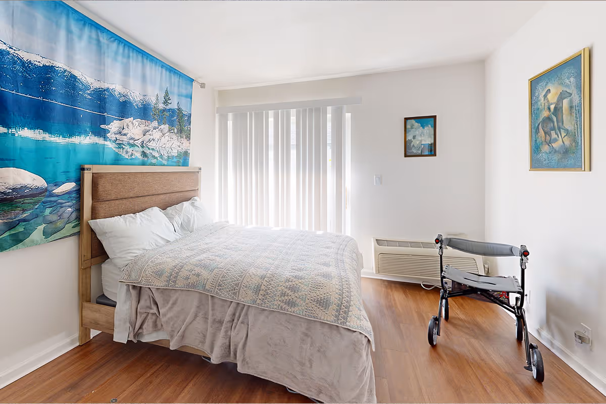 Sunlit bedroom with a bed against a wall featuring a lake tapestry, vertical blinds, framed art, and a rolling walker near the foot of the bed.