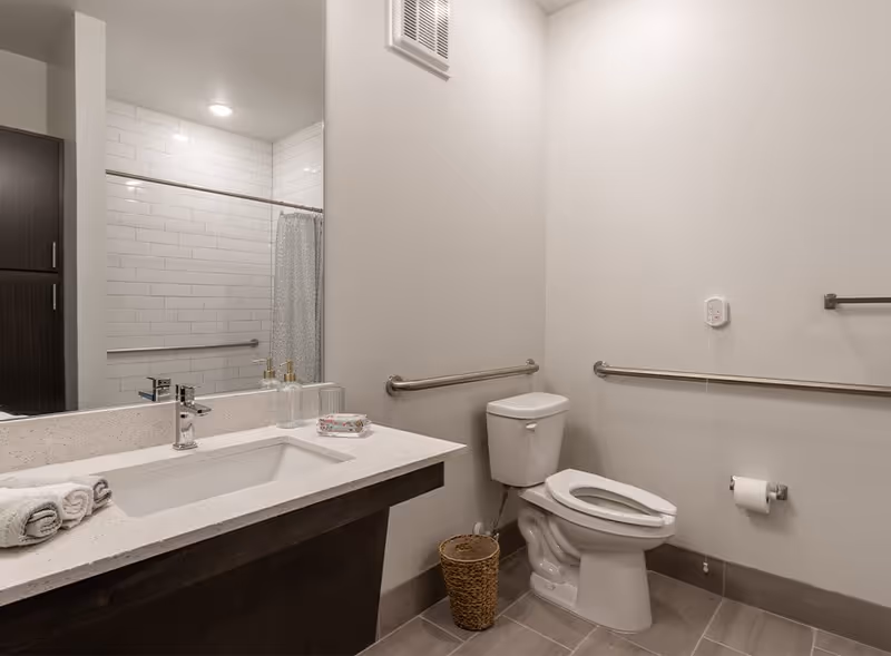A clean, modern bathroom with a white toilet, grab bars on the walls, a sink with a countertop, a large mirror, and a shower area with a curtain and white subway tile walls. There is a small wicker wastebasket next to the toilet and a folded towel on the sink counter.