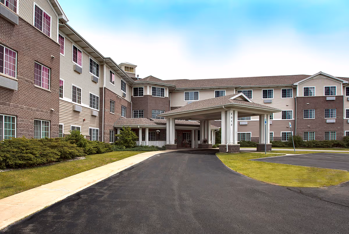 Front exterior view of a three-story senior living facility building with a covered entrance driveway, surrounded by green shrubs and a clear blue sky.
