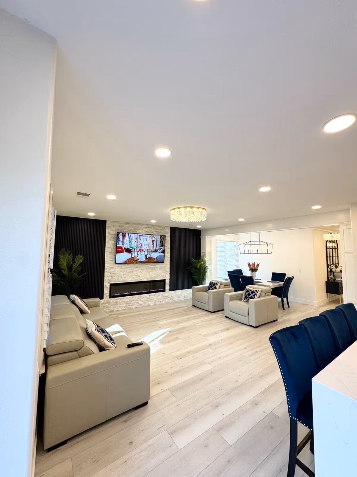 A modern senior living facility common area with light wood flooring, beige sofas and armchairs, a wall-mounted flat screen TV above a sleek electric fireplace, and a dining area with a white table and blue chairs. The room is well-lit with recessed ceiling lights and a decorative chandelier above the dining table. There are plants placed near the TV wall, and a kitchen counter with blue bar stools is partially visible on the right.