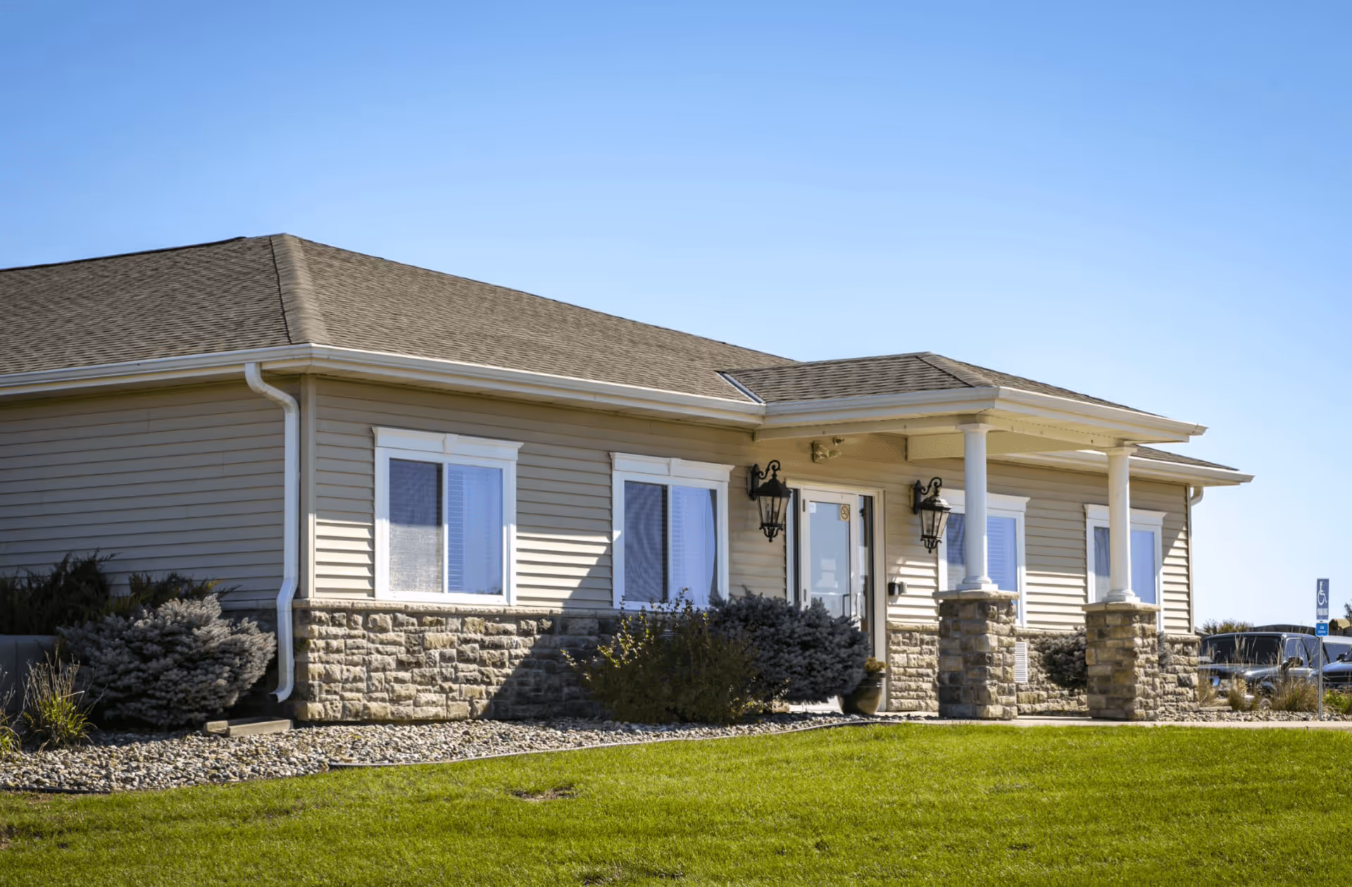 Exterior view of a single-story building with beige siding and stone accents, a small covered porch supported by white columns, and a well-maintained green lawn under a clear blue sky.