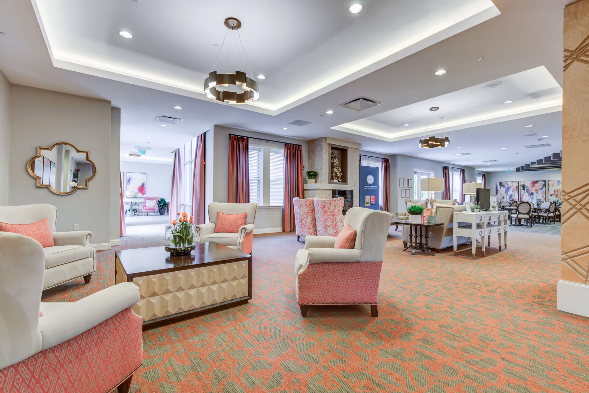 A spacious and well-lit senior living facility common area with comfortable armchairs featuring coral and beige upholstery, a wooden coffee table with a flower arrangement, and a patterned carpet in coral and beige tones. The room has large windows with coral curtains, modern ceiling lights, and a fireplace. In the background, there is a dining area with tables and chairs, and decorative artwork on the walls.
