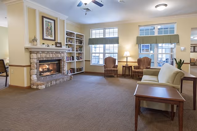 A cozy living room area with a stone fireplace on the left, built-in bookshelves, two armchairs with a small table and lamp between them near large windows with green valances, and a beige sofa with a wooden side table in the foreground. The room has beige walls, carpeted floor, and ceiling fans with lights.