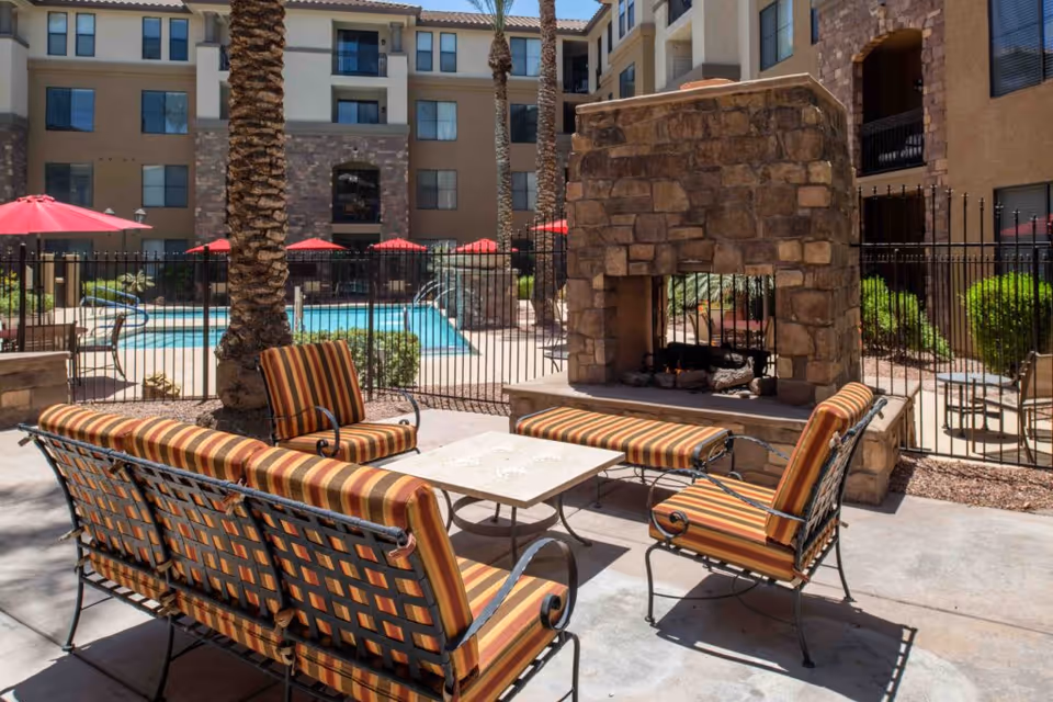 Outdoor seating area with striped cushioned chairs and a stone fireplace, surrounded by palm trees and a fenced swimming pool with red umbrellas in the background, located within a multi-story residential building complex.