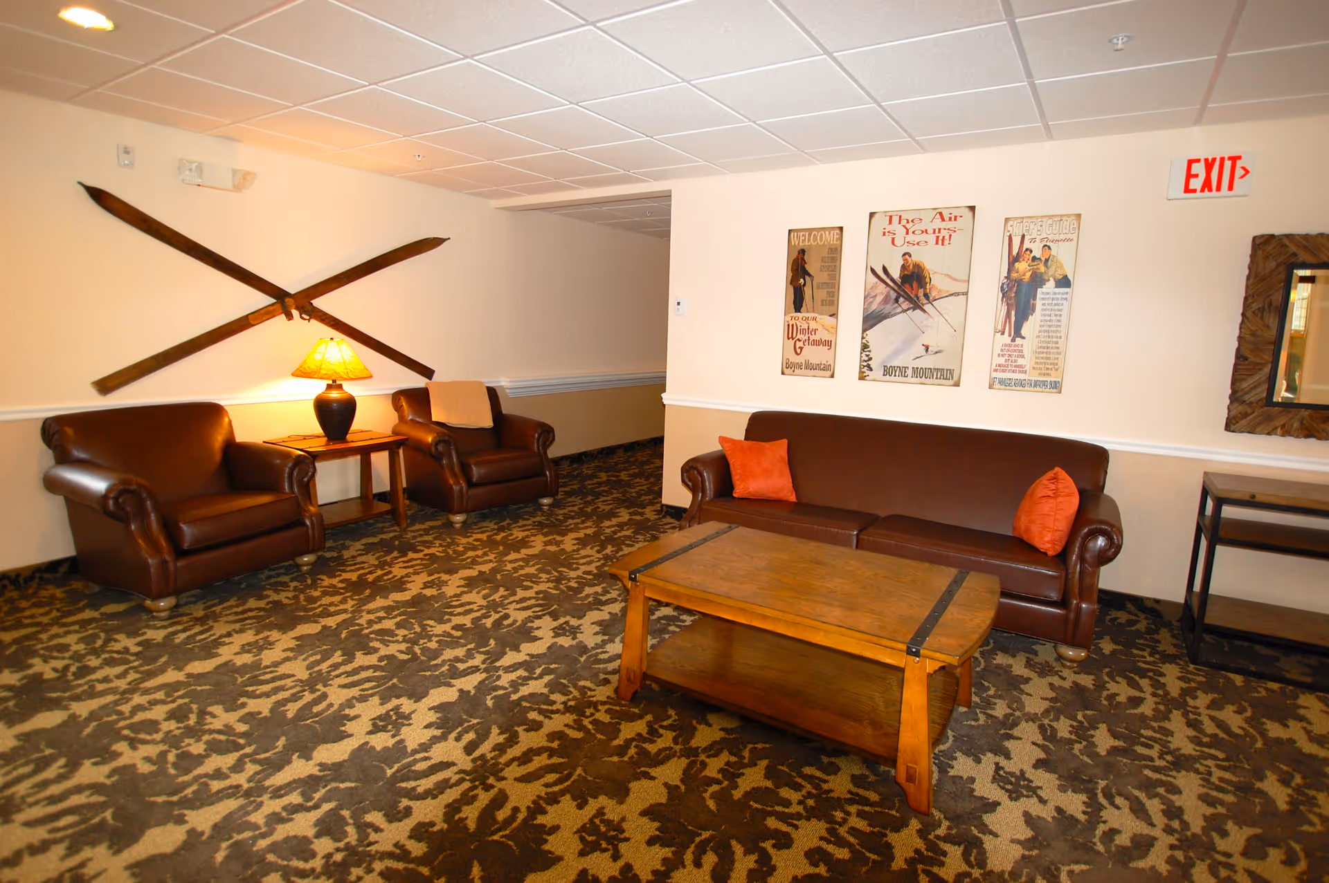 A cozy living room area with a brown leather sofa and two matching armchairs. There is a wooden coffee table in front of the sofa and a side table with a lamp between the armchairs. The walls are decorated with vintage ski-themed posters and crossed wooden skis. The carpet has a leafy pattern, and there is an exit sign above a doorway on the right.