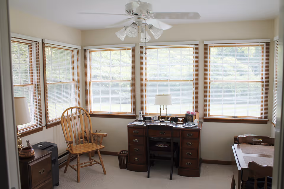 A bright room with multiple large windows covered with wooden blinds. The room contains a wooden desk with a lamp, telephone, calculator, and various office supplies. There is a wooden chair in front of the desk and another wooden chair to the left near a small side table with a lamp. A ceiling fan with lights is mounted on the ceiling.
