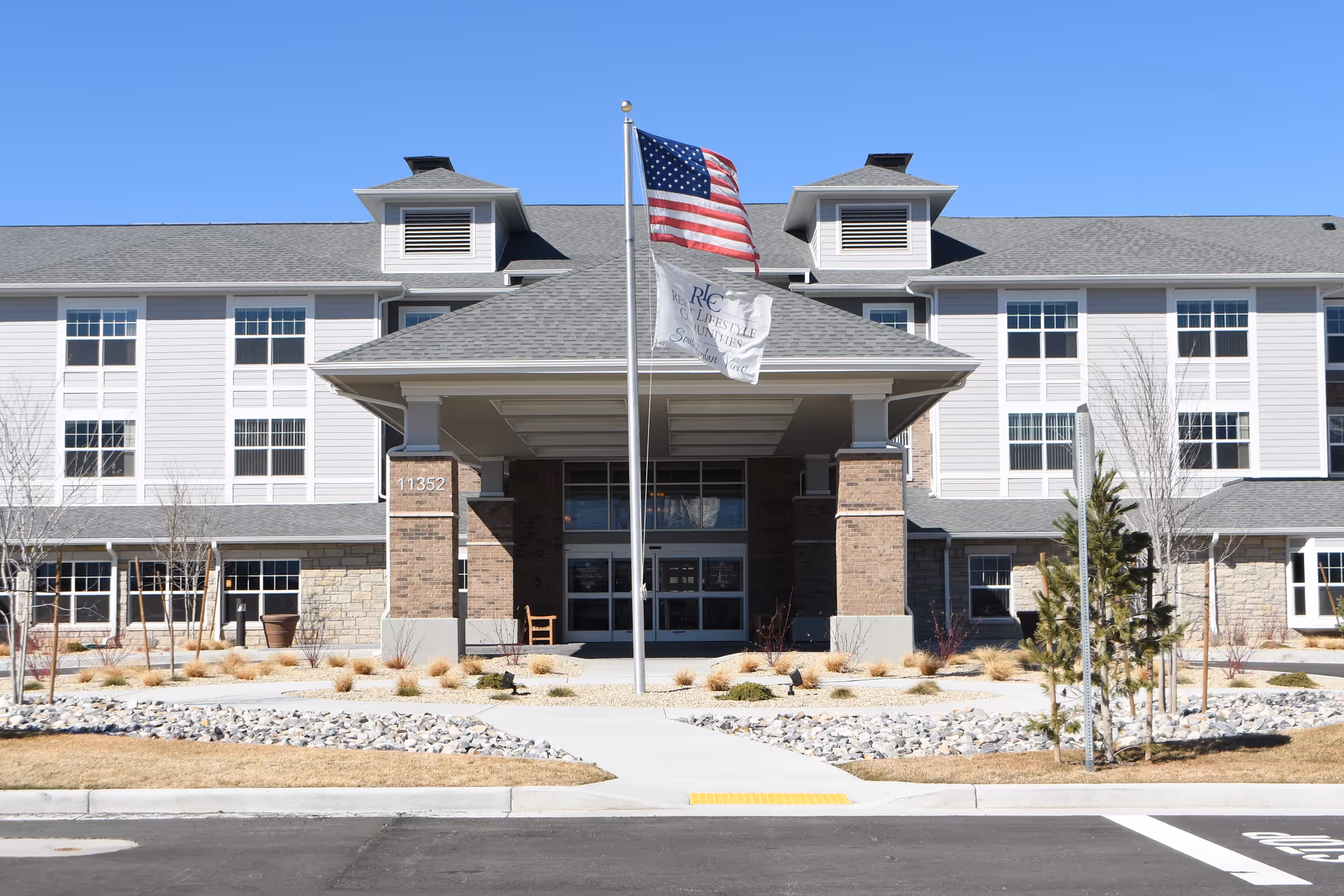 Front exterior view of South Jordan View Retirement Community building with an American flag and a community flag flying on a flagpole in front. The building has multiple windows, a covered entrance supported by brick columns, and landscaping with rocks and small plants.