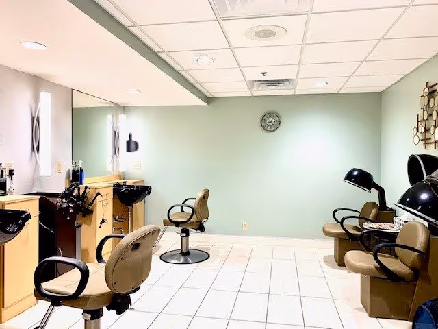Interior of a hair salon area in a senior living facility with salon chairs, hair washing sinks, mirrors, and hair drying stations against light green and beige walls with a clock on the wall.