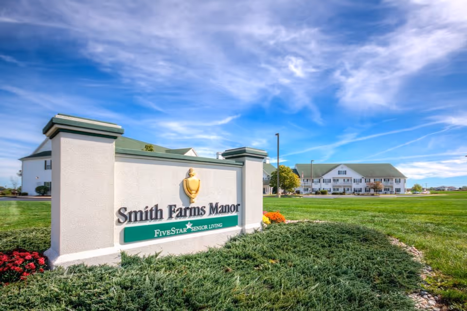 Exterior view of Smith Farms Manor senior living facility with a large sign in the foreground displaying the facility name and Five Star Senior Living logo, surrounded by well-maintained landscaping and a bright blue sky with scattered clouds.