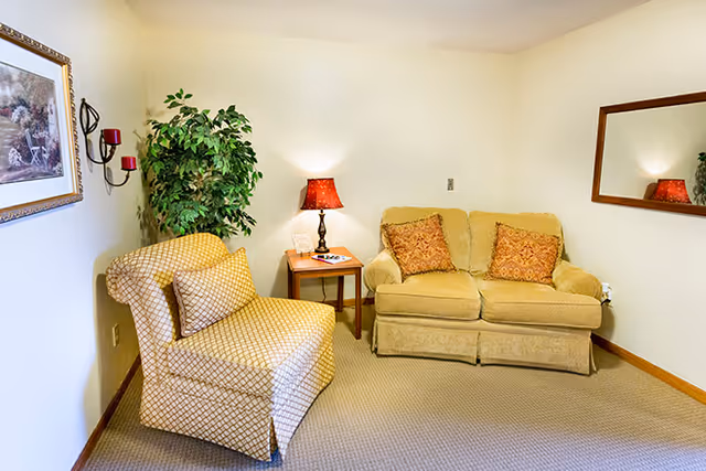 Cozy sitting area with a yellow loveseat and patterned armchair flanking a side table and lamp against a neutral wall.