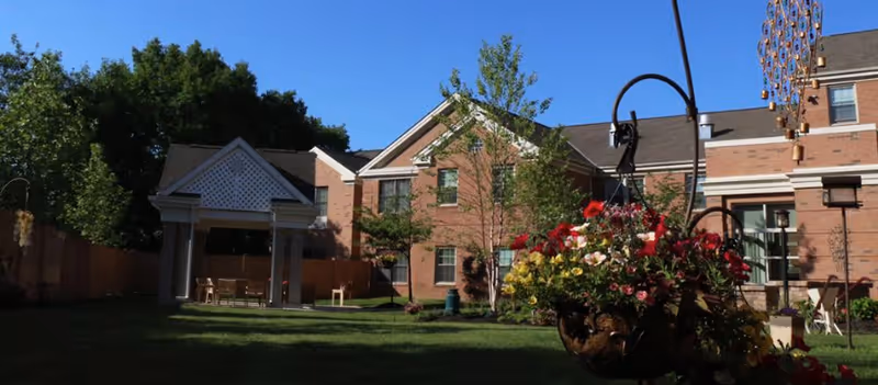 Outdoor garden area of a senior living facility with green grass, trees, a hanging flower basket with red and yellow flowers, a covered seating area with chairs and tables, and a brick building in the background under a clear blue sky.