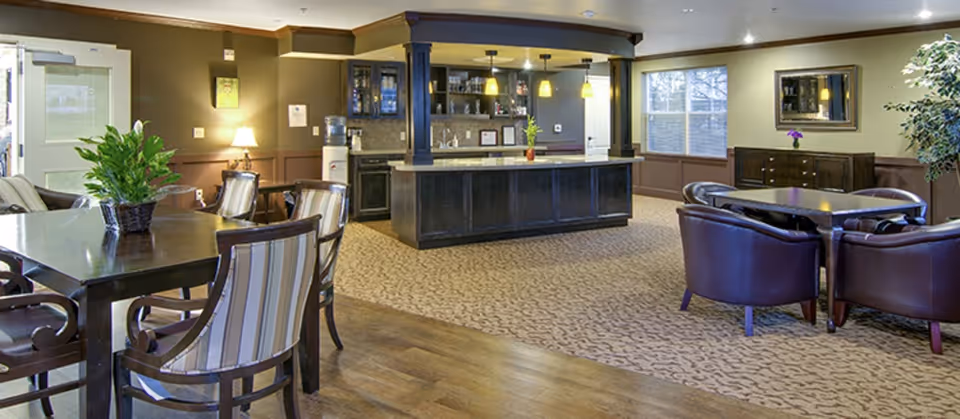 A spacious common area with a combination of carpet and wood flooring, featuring multiple seating arrangements including a table with striped chairs and another table with leather armchairs. There is a central counter with cabinets and a sink, pendant lights hanging above, a water cooler, and a window letting in natural light. The walls are painted in warm tones with decorative molding and a mirror above a sideboard.