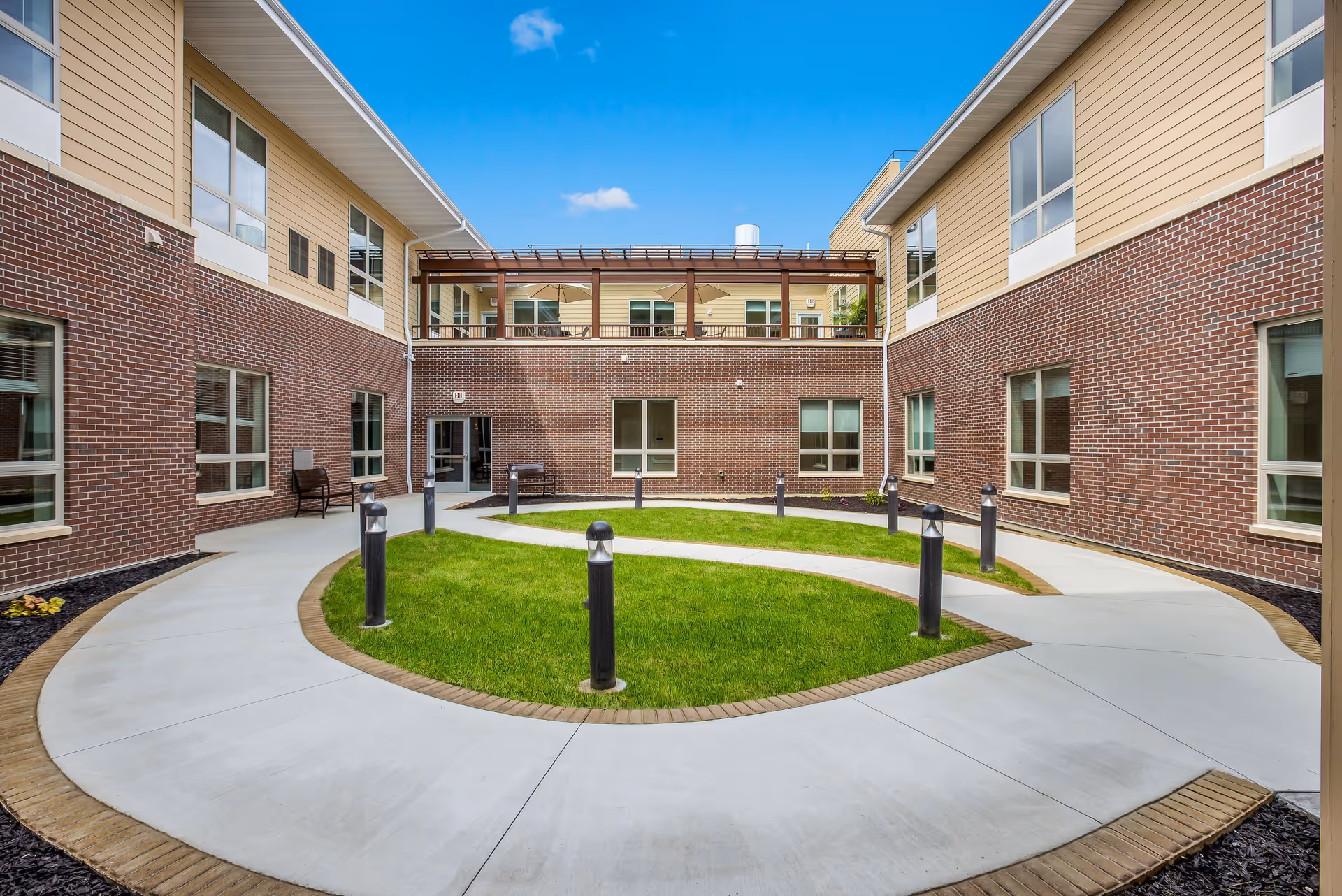 Outdoor courtyard area at Cascade Creek facility featuring a curved concrete walkway surrounding a green grassy area with black bollard lights. The courtyard is enclosed by a two-story building with brick and beige siding exterior walls and multiple windows. There is a covered balcony on the second floor and a bench near a door on the ground level. The sky is clear and blue.