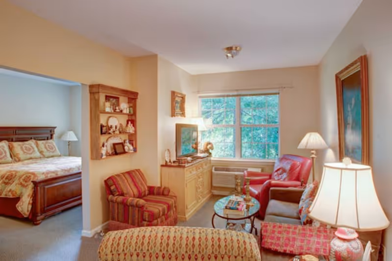 Cozy assisted-living suite with a living area featuring armchairs, a sofa, coffee table and lamps, and a bedroom visible through an open doorway.
