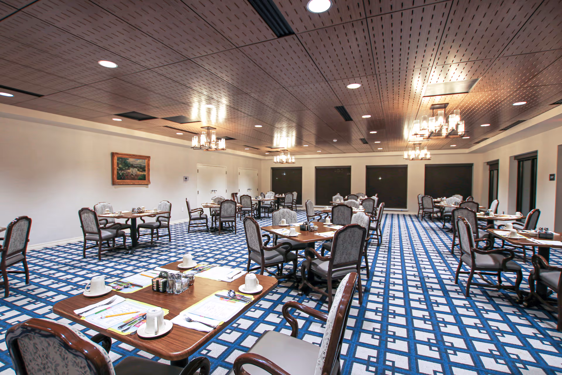 A spacious dining room with multiple wooden tables and cushioned chairs arranged neatly. Each table is set with white cups, saucers, utensils, and napkins. The room features a patterned blue and white carpet, a wooden paneled ceiling with recessed lighting, and several chandeliers. There are windows with dark blinds and a framed painting on one wall.