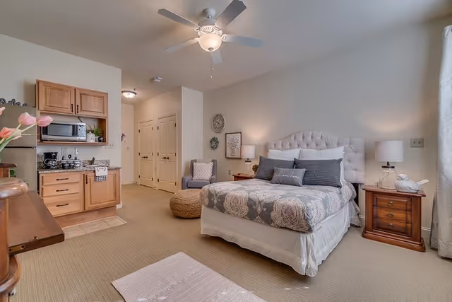 A cozy bedroom with a queen-sized bed featuring a tufted headboard and floral bedding. There are two wooden nightstands with lamps on either side of the bed. A comfortable armchair and a round woven ottoman are placed near the bed. To the left, there is a small kitchenette with wooden cabinets, a microwave, coffee maker, and a stainless steel refrigerator. The room has beige carpeting, light-colored walls, and a ceiling fan with a light fixture.