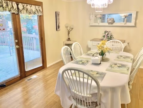 A dining room with a rectangular table covered with a white tablecloth and six white chairs around it. The table is set with floral placemats and a centerpiece with yellow flowers. There is a sideboard against the wall with decorative items and a large mirror above it. French doors with blinds lead to an outdoor deck, and the room has light-colored walls and wooden flooring.