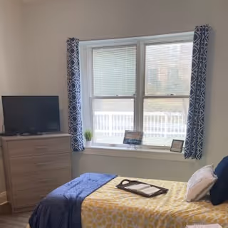A bedroom with a bed covered in a yellow and white patterned blanket and a blue throw blanket. There are pillows on the bed, and a tray with a book and glasses rests on the bed. A wooden dresser with a flat-screen TV on top is positioned next to a window with blue patterned curtains. The window has blinds and overlooks an outdoor area with a white fence.
