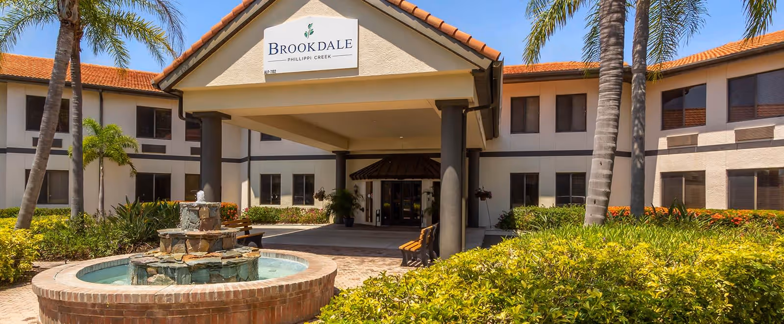 Exterior view of Brookdale Phillippi Creek senior living facility with a covered entrance, a brick water fountain in the foreground, palm trees, and well-maintained landscaping under a clear blue sky.