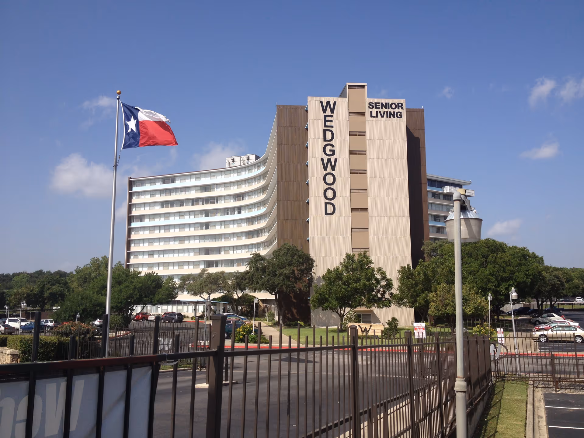 Exterior view of Wedgwood Senior Apartments, a multi-story senior living building with a curved facade and large windows. A Texas state flag flies on a flagpole in front of the building, and there are trees, parked cars, and a gated entrance visible in the foreground under a clear blue sky.