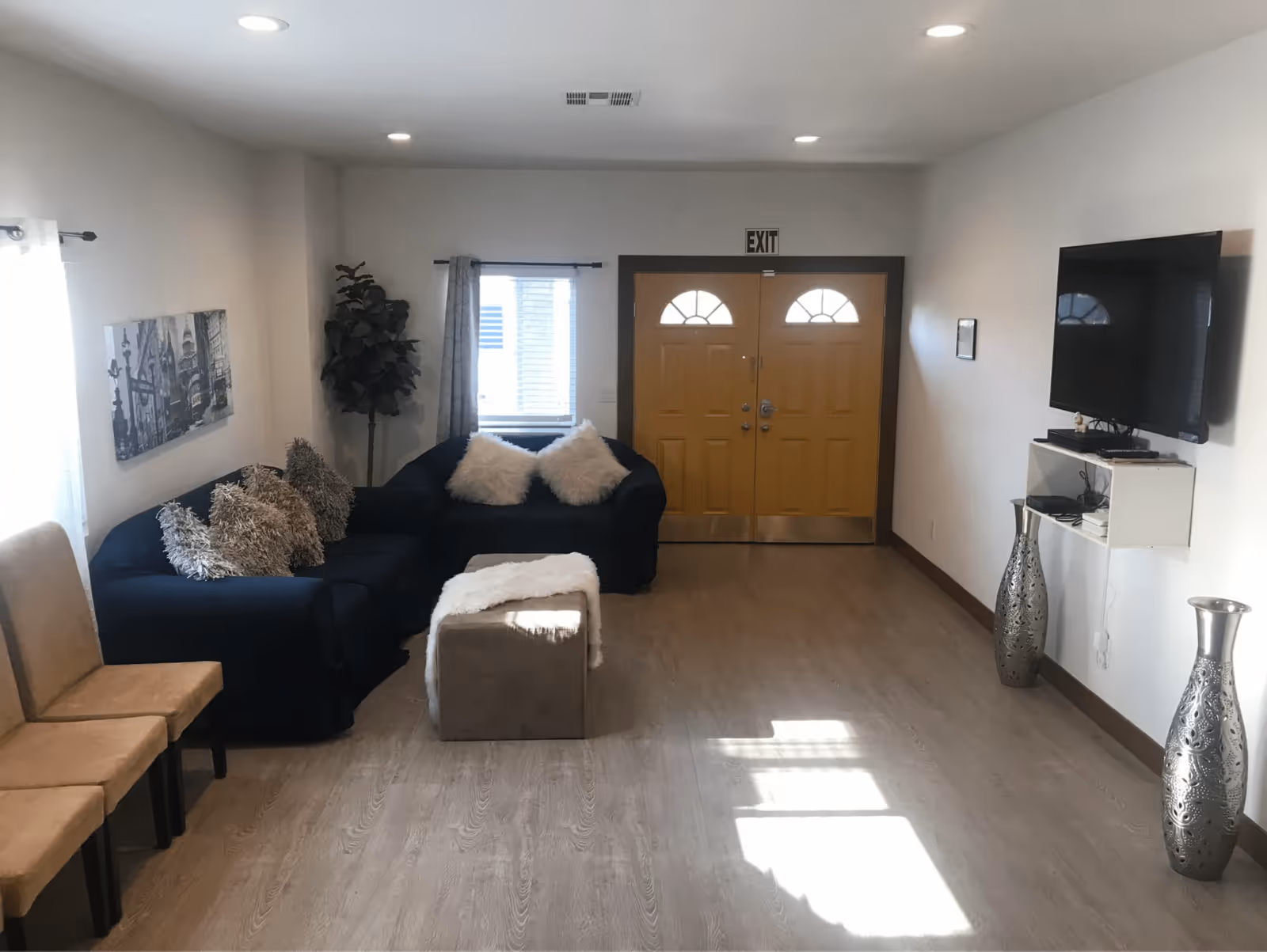 A bright living room with light wood flooring, a dark blue sectional sofa adorned with fluffy pillows, a small ottoman with a white throw, two beige chairs, a wall-mounted flat-screen TV, two tall decorative silver vases, a window with curtains, and double wooden doors with an exit sign above.