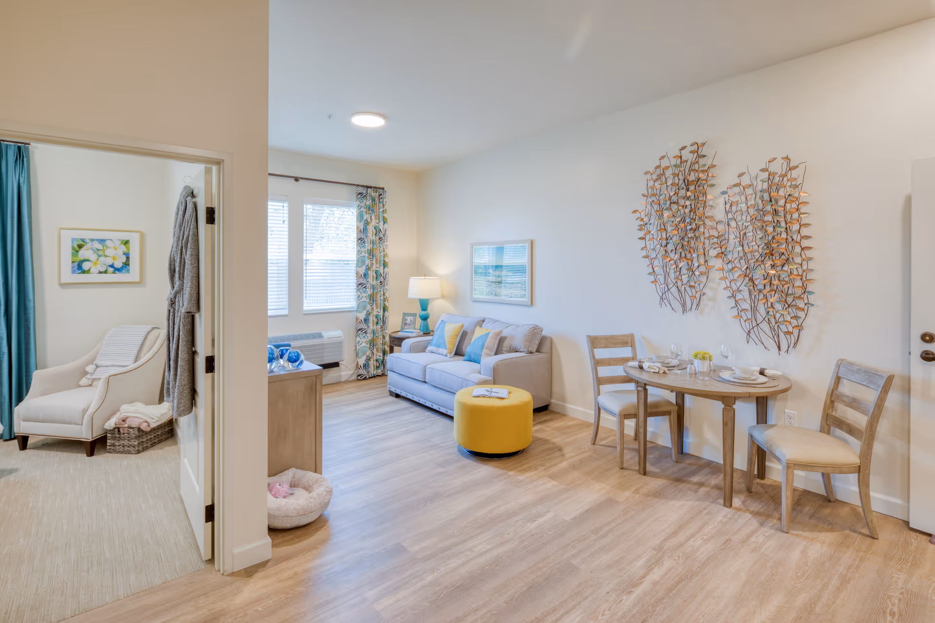 A bright and cozy living room area with a gray sofa adorned with colorful pillows, a yellow ottoman, a small wooden dining table set with two chairs, and decorative metal wall art. To the left, an open doorway leads to a room with a beige armchair, a basket with folded blankets, and a framed floral picture on the wall. The space has light wood flooring and large windows with patterned curtains letting in natural light.