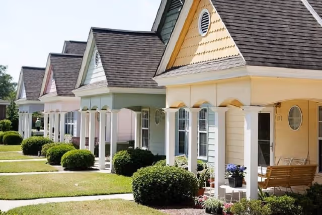 Row of small, colorful cottages with front porches, each with white columns and neatly trimmed bushes in front, under clear daylight.