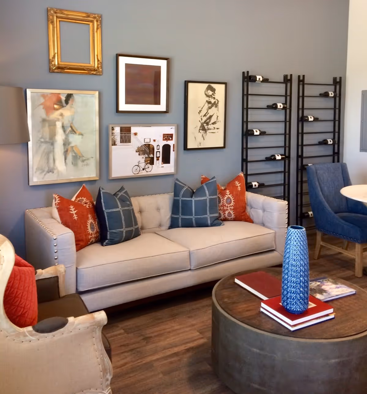 A cozy living room area with a beige tufted sofa adorned with blue and orange decorative pillows. On the wall behind the sofa are four framed artworks, including an empty gold frame, a painting of a woman, a photo of a bicycle in front of a building, and a sketch of a person. To the right, there are two black wine racks with several bottles of wine. In front of the sofa is a round wooden coffee table with a tall blue vase and some books. A beige armchair with a red cushion and a blue upholstered chair are also visible.