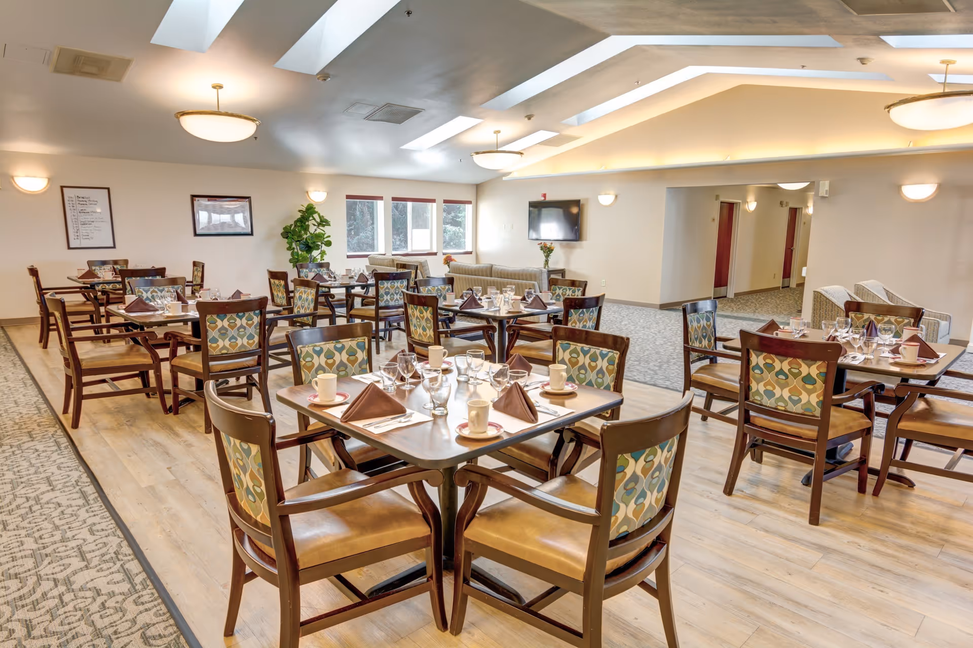 A bright and spacious dining room with multiple wooden tables and chairs arranged neatly. Each table is set with cups, glasses, napkins, and utensils. The room features skylights, ceiling lights, and large windows allowing natural light to enter. There are some couches and plants in the background, and the floor is a combination of wood and carpet.