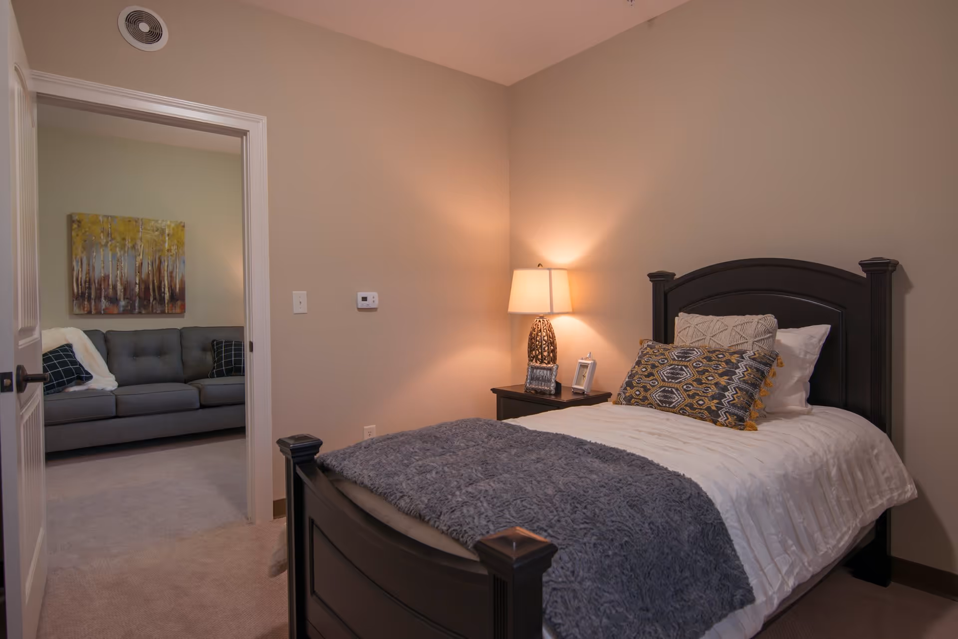 A cozy bedroom with a dark wooden bed frame, white bedding, and decorative pillows. A nightstand with a lamp and picture frames is beside the bed. The door is open, showing a glimpse of an adjacent room with a gray sofa and a painting of trees on the wall.