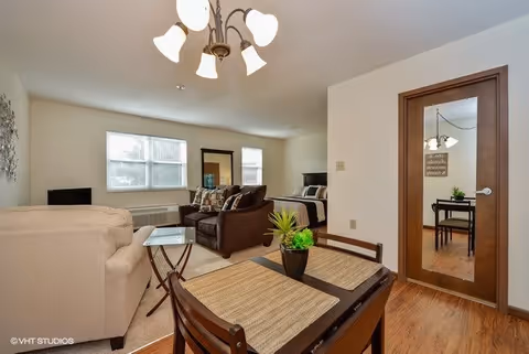 A cozy living area with a beige armchair and a brown sofa arranged around a glass coffee table. Behind the sofa, there is a bed visible in a separate sleeping area. A small dining table with two chairs and placemats is in the foreground, with a small potted plant on it. A door with a glass panel reflects part of the room, and two windows allow natural light to brighten the space. The room has wooden flooring and a ceiling light fixture with multiple bulbs.