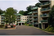 Exterior view of a multi-story senior living facility with balconies, surrounded by greenery and trees, with a driveway and parked cars in front.