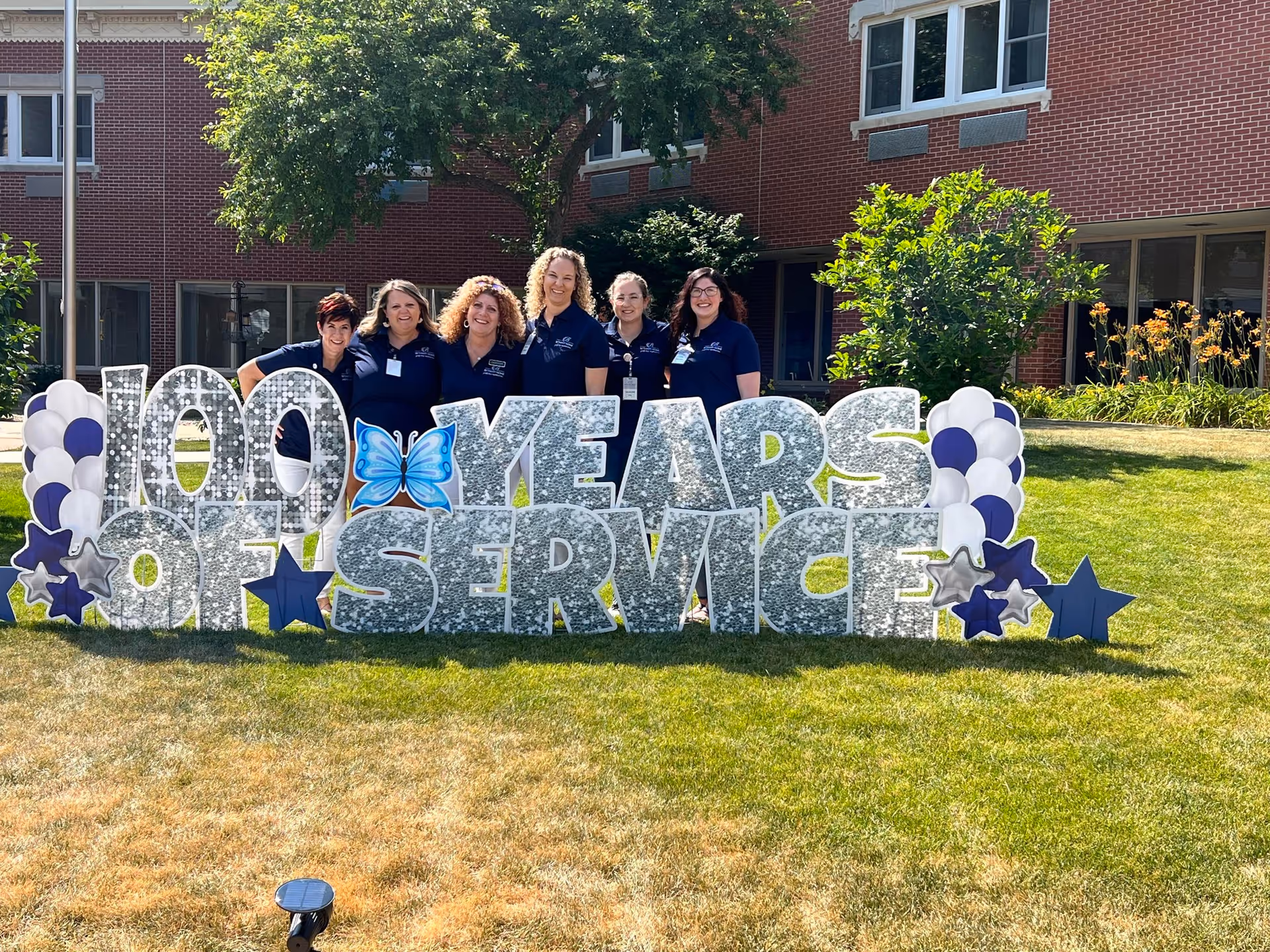 Six staff members stand behind a large decorative "100 YEARS OF SERVICE" sign on the lawn in front of a brick retirement center.