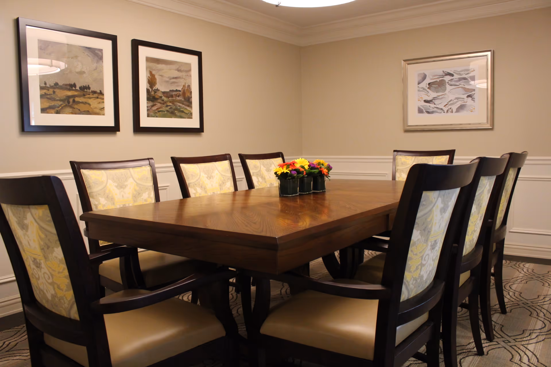 A dining room with a large wooden table surrounded by upholstered chairs and a small floral centerpiece.