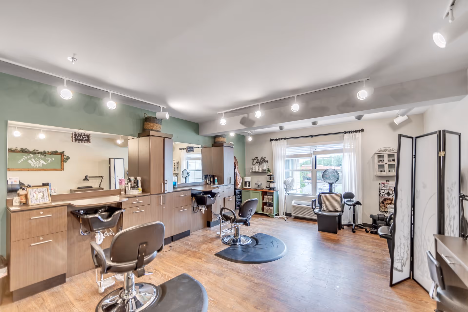 Interior of a bright and clean hair salon area with wooden flooring, multiple styling chairs, large mirrors, and cabinets. The room has a green accent wall with track lighting on the ceiling, a window with sheer white curtains, and various hair care products and equipment neatly arranged.