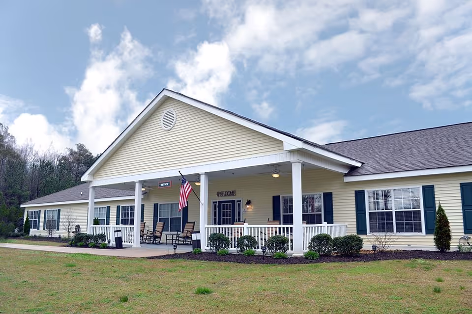 Exterior view of a single-story yellow building with white trim and a covered porch. The porch has rocking chairs, an American flag, and a sign that says 'Office' above the entrance. The building is surrounded by grass and some shrubs under a partly cloudy sky.