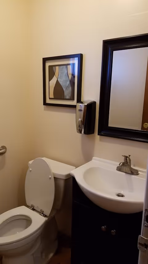 A small bathroom with a white toilet with the lid open, a white sink with a silver faucet, a dark-framed mirror above the sink, a wall-mounted soap dispenser, and a framed abstract artwork on the wall.