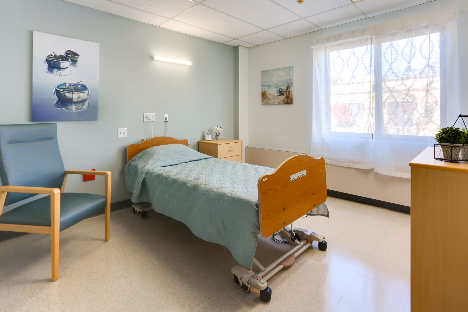 A simple, clean bedroom in a senior living facility featuring a single hospital-style bed with a light blue quilt, a blue armchair with wooden arms, a small wooden dresser, and two paintings on the walls. A large window with sheer white curtains allows natural light to fill the room.