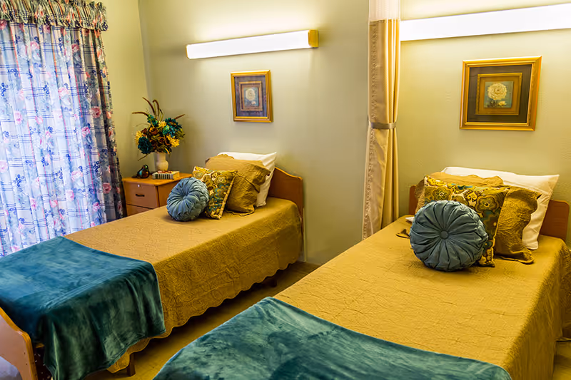 A cozy nursing facility bedroom with two single beds, each covered with a mustard yellow bedspread and a teal throw blanket at the foot. Each bed has decorative pillows including a round teal pillow and patterned cushions. Between the beds is a small wooden nightstand with a vase of flowers and a telephone. The room has floral curtains on the window and framed artwork on the walls above each bed. A long fluorescent light fixture is mounted on the wall above the beds.