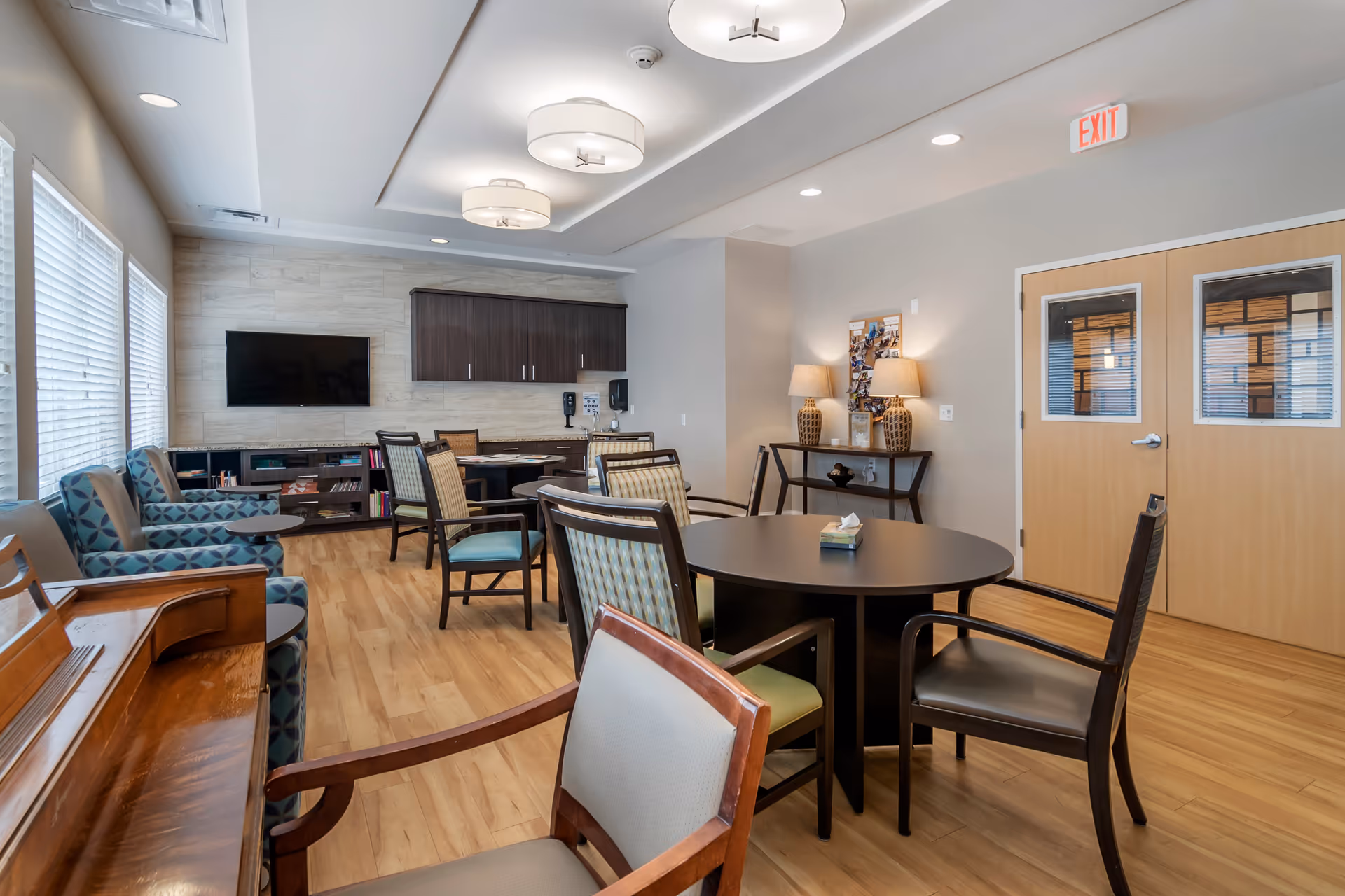 A well-lit common area in a senior living facility featuring multiple chairs around round tables, a piano, a wall-mounted TV, cabinets, and two table lamps on a console table near double doors with windows. The room has large windows with blinds and wood flooring.