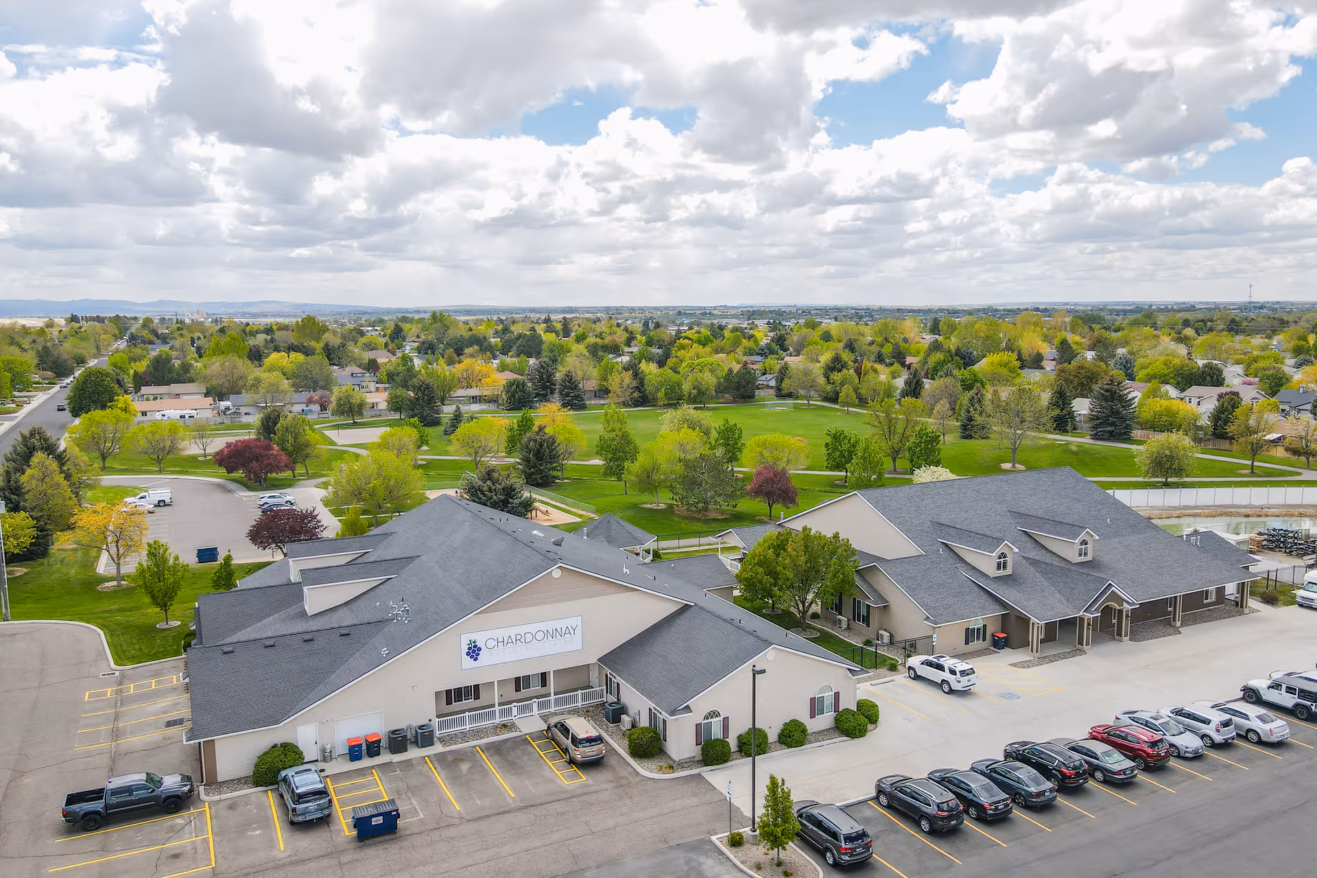 Aerial view of the Chardonnay Assisted Living building with a parking lot in front and a green park and neighborhood behind it.