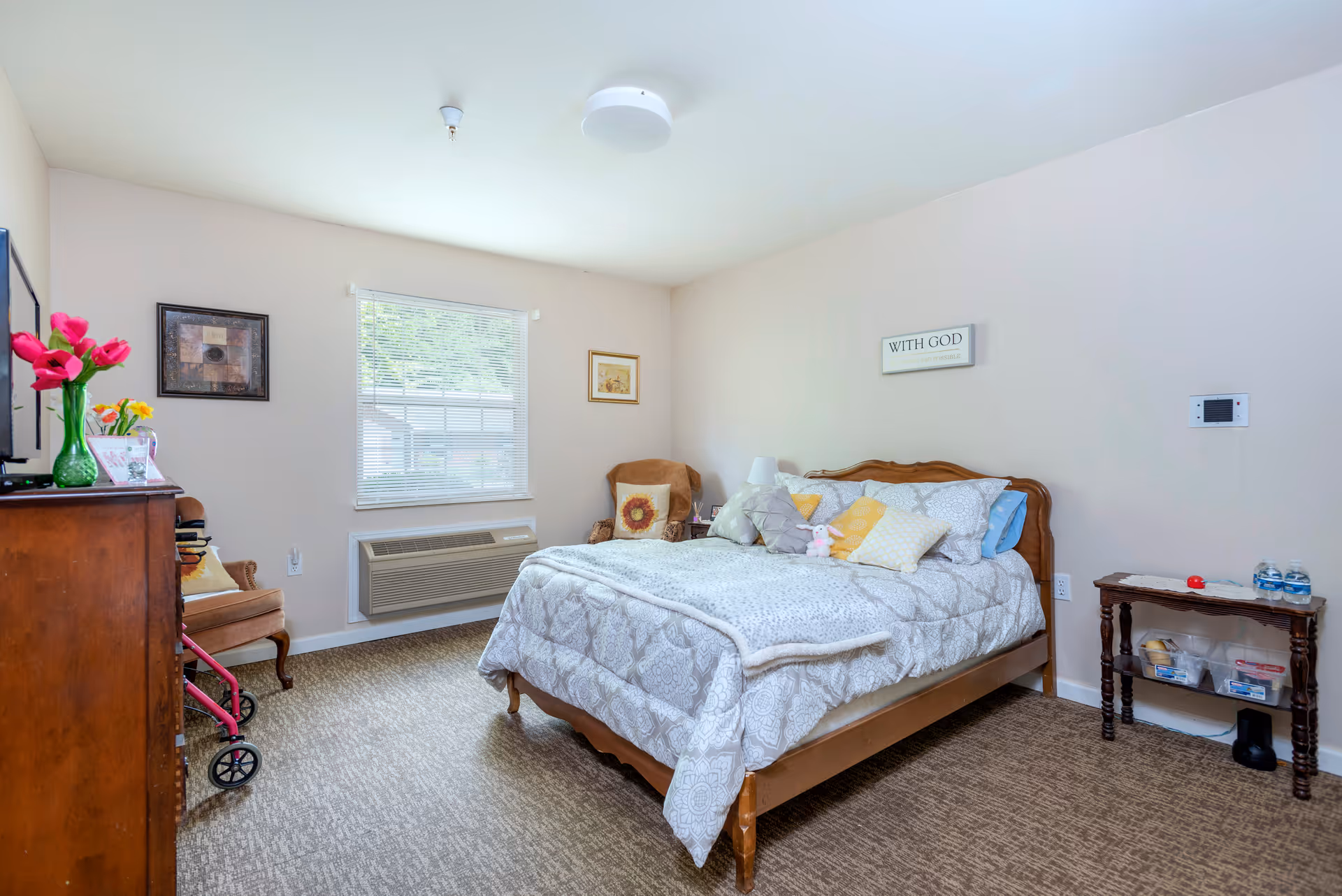 A cozy bedroom in a senior living facility with a neatly made bed featuring multiple pillows and a stuffed animal. The room has a window with blinds, a wooden dresser with a vase of flowers, a chair with a cushion, and a small table holding water bottles and other items. The walls are light-colored with framed artwork and a sign that reads 'WITH GOD'.